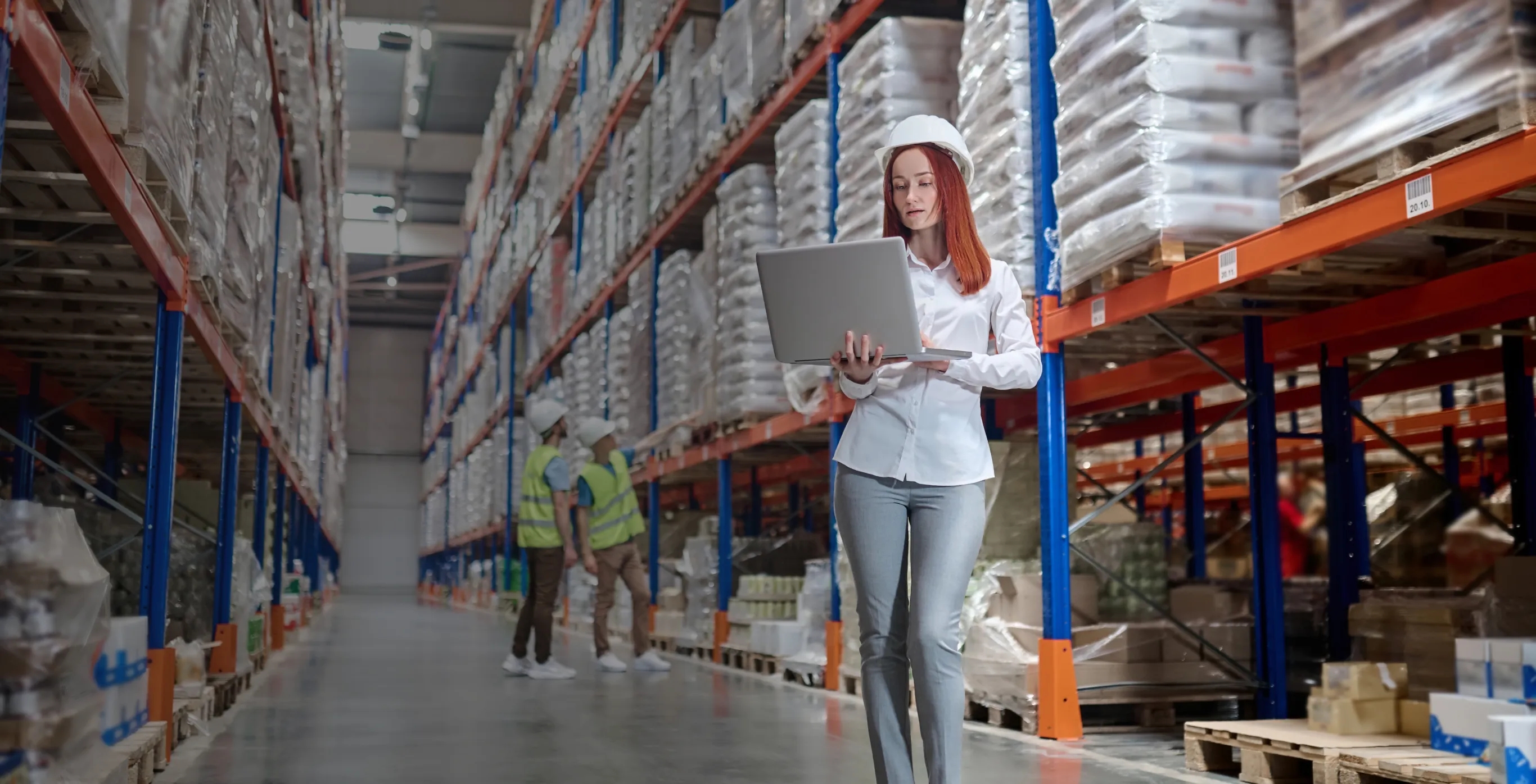 A woman in a white shirt and hat stands in a warehouse, working on a laptop, representing tech solutions for logistics.